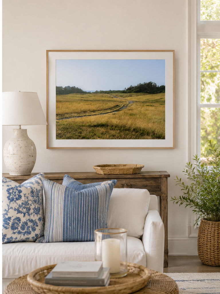 A framed landscape art print depicting a grassy field with a winding path under a blue sky, styled in a bright living room above a white sofa with blue patterned pillows.